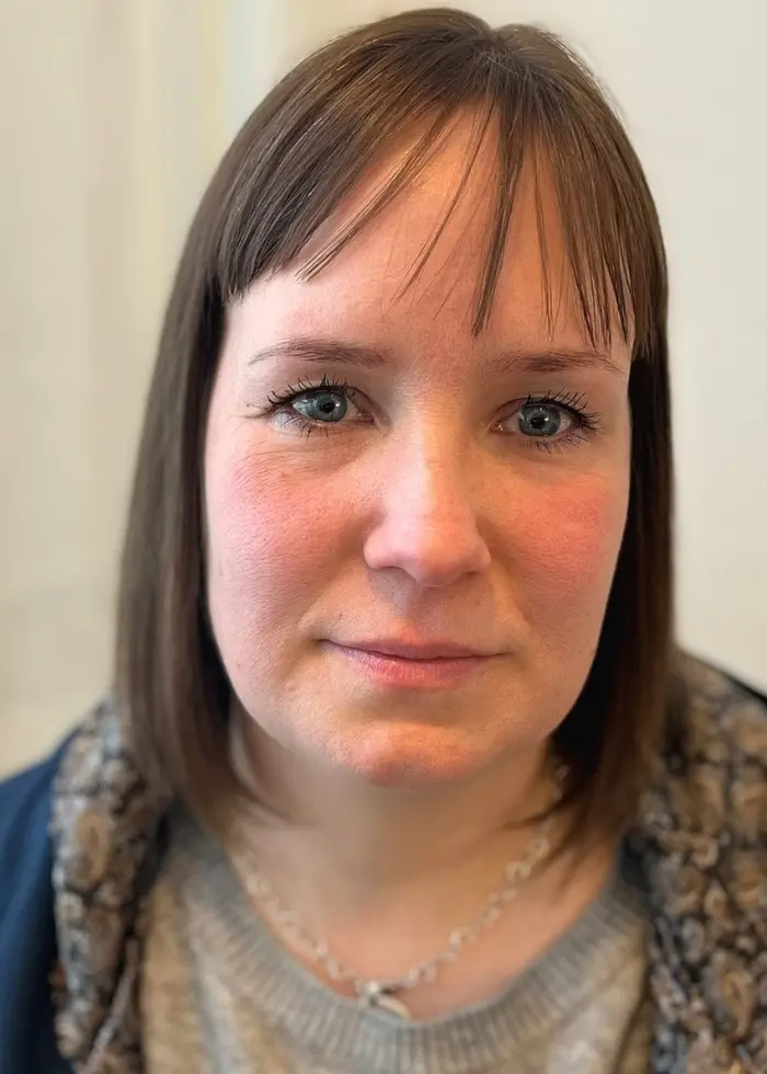 En person med axellångt hår står framför ett vitt draperi. Personen bär en grå tröja och en mörk jacka med foder. Bilden är tagen inomhus.