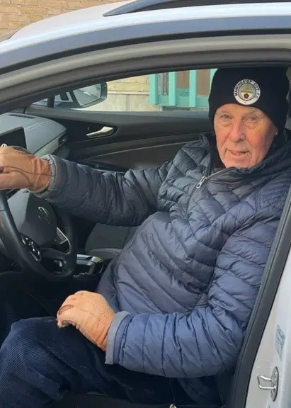 En person sitter i förarsätet på en vit elbil med dörren öppen. Personen bär en mörk jacka och keps. I bakgrunden syns en gul byggnad med stora fönster.