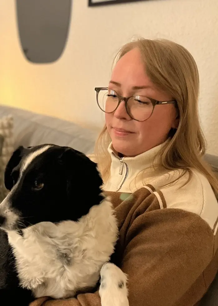 En person sitter i en soffa med en svartvit hund i knät. Bakgrunden visar en ljus vägg med tavlor och en grön växt. Soffan är dekorerad med kuddar i beige och gröna toner.