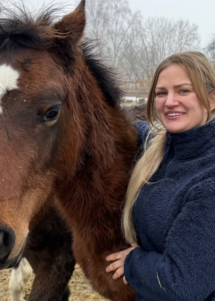 En person klädd i en blå tröja står nära två hästar på en snötäckt gård med träd i bakgrunden. Hästarna är bruna och har vita tecken i ansiktet.