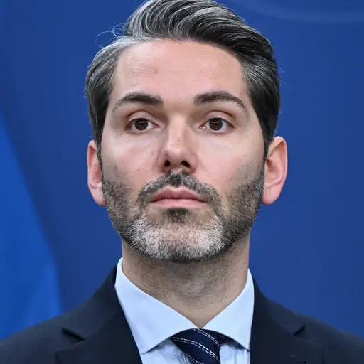 En man i kostym och slips står framför en blå bakgrund med delar av en svensk flagga synlig. Han deltar i en pressträff om migration och socialförsäkringspolitik.