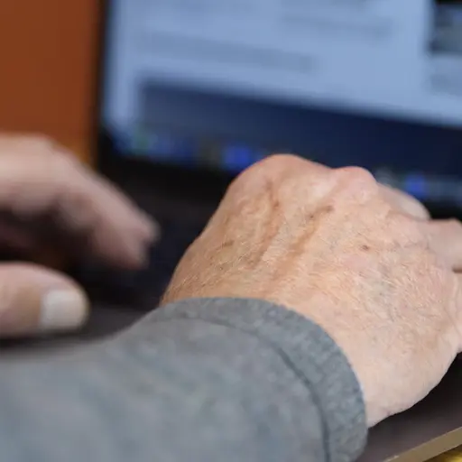 En äldre man använder en bärbar dator vid ett bord. Bilden visar hans händer på tangentbordet, med en skärm i bakgrunden som visar olika webbplatser och annonser.