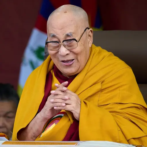 Den tibetanske andlige ledaren Dalai Lama sitter vid ett bord klädd i en gul dräkt. Bordet är dekorerat med en färgglad duk och en orkidé står bredvid. Flera flaggor syns i bakgrunden.