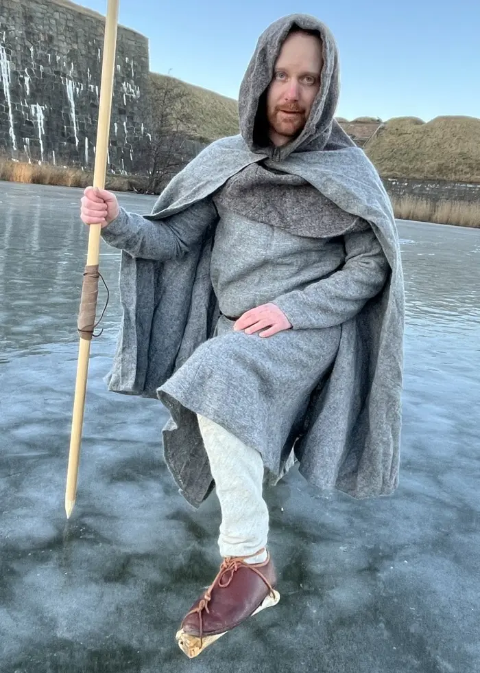 En person klädd i en grå mantel står på en frusen vallgrav med en stav i handen. Personen är omgiven av ett vinterlandskap med fästning och klar himmel bakom.