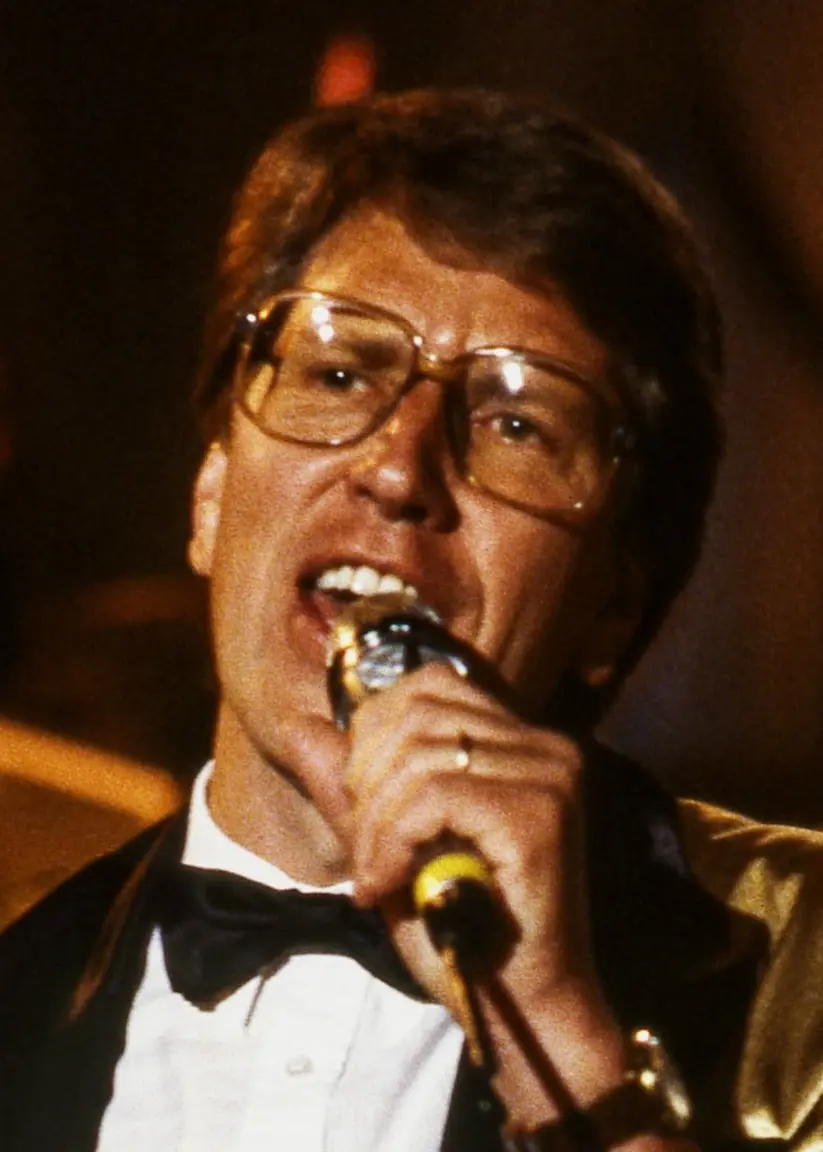 Stefan Borsch håller en mikrofon nära munnen med vänster hand. Han sjunger förmodligen. Han är kortklippt och bär glasögon.