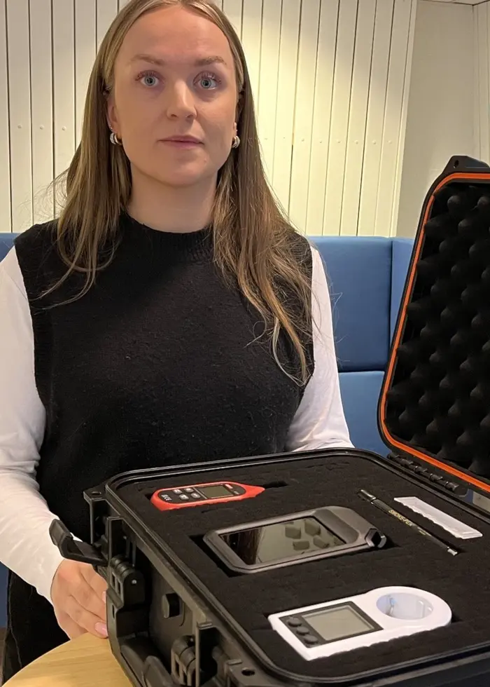 En person står vid ett bord med en svart väska med teknisk utrustning. Bakgrunden visar en blå soffa.