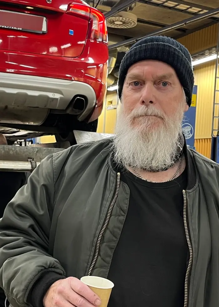 En man står i en bilverkstad med en röd bil upphissad på en lyft i bakgrunden. Han håller en kaffemugg i handen och är klädd i mörka kläder. Verkstadens väggar är gula.