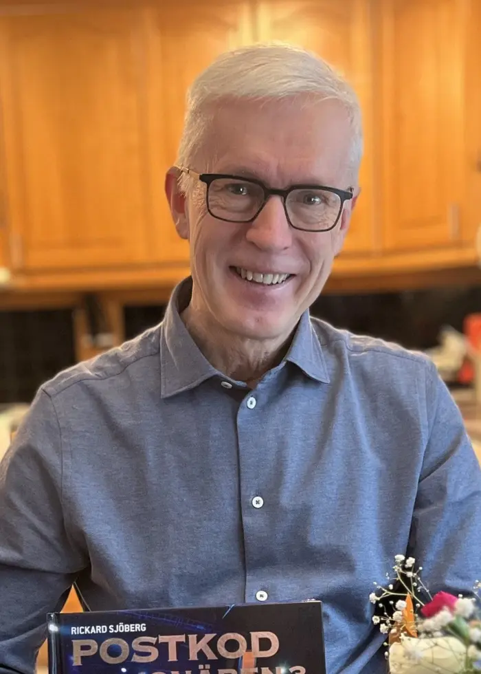 En person sitter i ett kök med träskåp och håller upp ett postkodlotteri-kuvert. På bordet framför personen finns en vas med blommor.