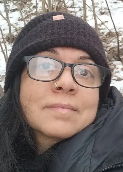 En person klädd i en mörk jacka och mössa står i en vinterskog med snö på marken och träd i bakgrunden.