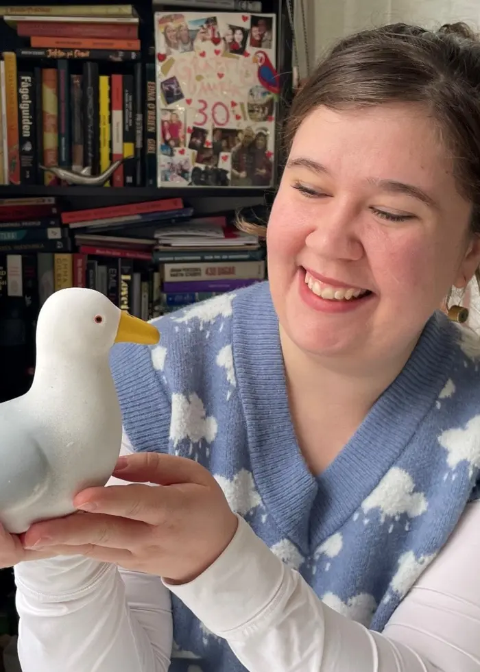 En person i en blå stickad väst med vita molnmönster håller en vitmålad prydnadsmås i händerna. Bakgrunden visar en bokhylla och ett fönster med vita gardiner.