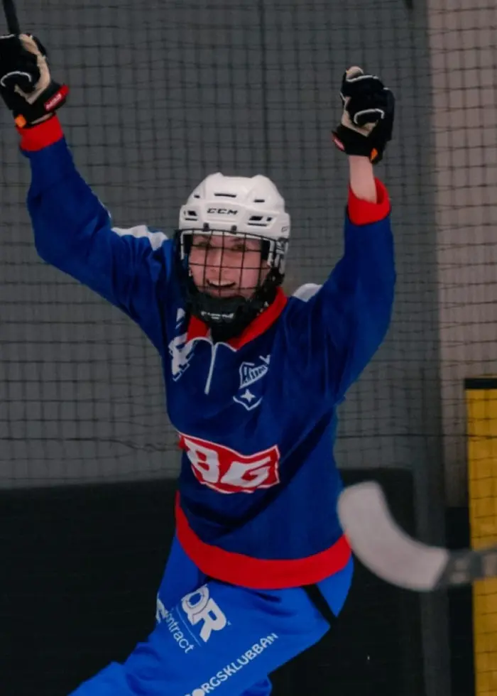 En bandyspelare i blå matchtröja firar ett mål med uppsträckta armar på en isplan. I bakgrunden syns ett blått reklamskylt och en motståndare i vit tröja med klubba.