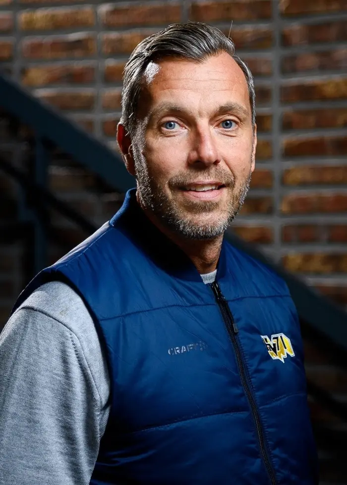 En person står framför en tegelvägg vid en trappa, klädd i en blå väst med en logotyp och en grå tröja. Miljön är mörkt belyst med fokus på personen och bakgrunden består av tegel och metallräcken.Hu