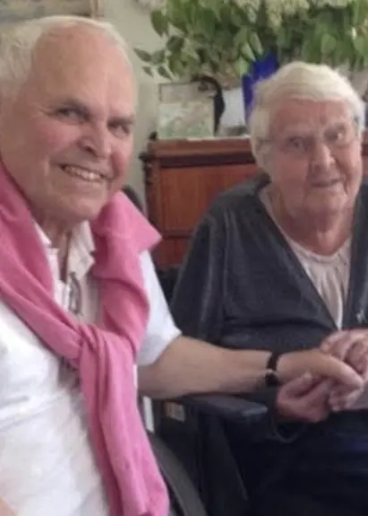 Två äldre personer sitter nära varandra i ett ljust rum med växter och möbler i bakgrunden. Den ena har en rosa tröja över axlarna och håller den andras hand.