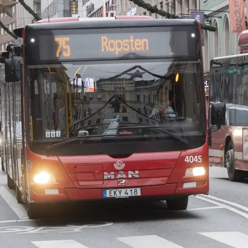 Två röda SL-bussar kör på en gata i centrala Stockholm. En person med gul mössa står vid ett övergångsställe och väntar på att korsa gatan. Stadsmiljö med byggnader och dekorationer syns i bakgrunden.