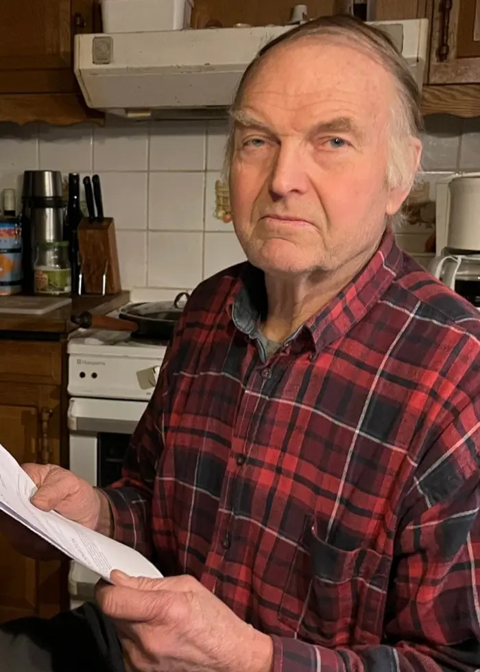 En man i rutig skjorta sitter i ett kök med träinredning och håller ett papper i handen. Köksbänken är fylld med burkar och andra köksartiklar.