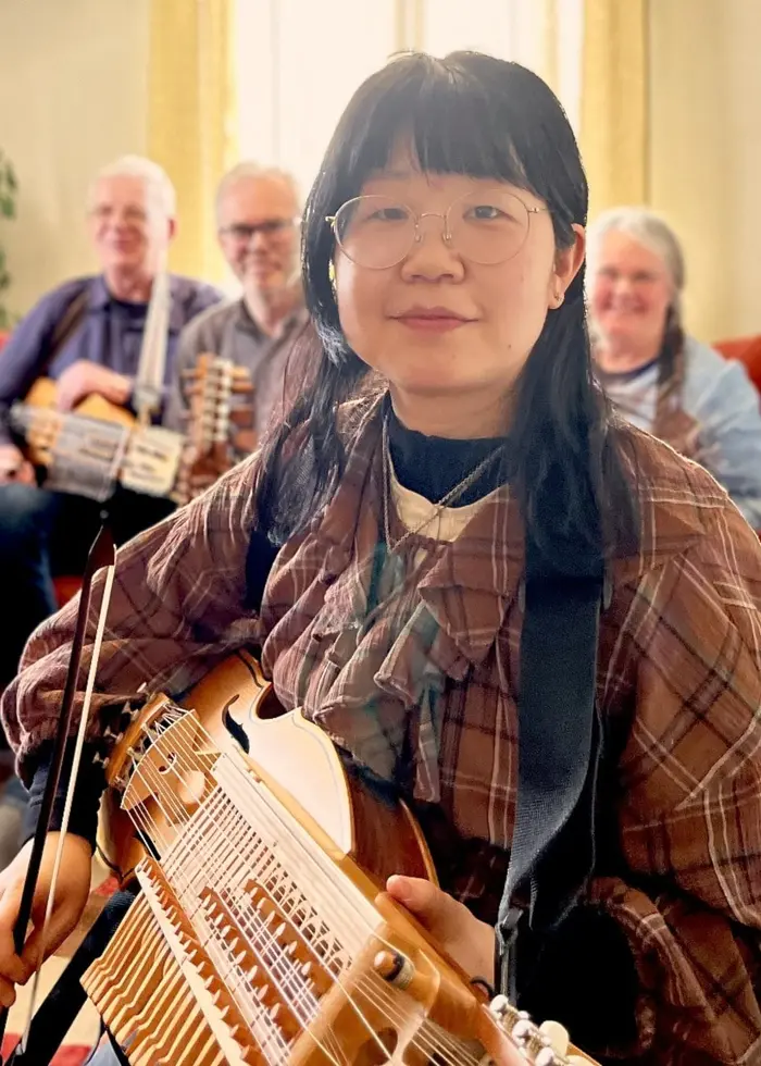 En grupp människor sitter tillsammans i ett vardagsrum och spelar nyckelharpa. En person i förgrunden håller i en nyckelharpa, medan andra i bakgrunden också spelar instrument. Rummet är ljust och hemtrevligt inrett.