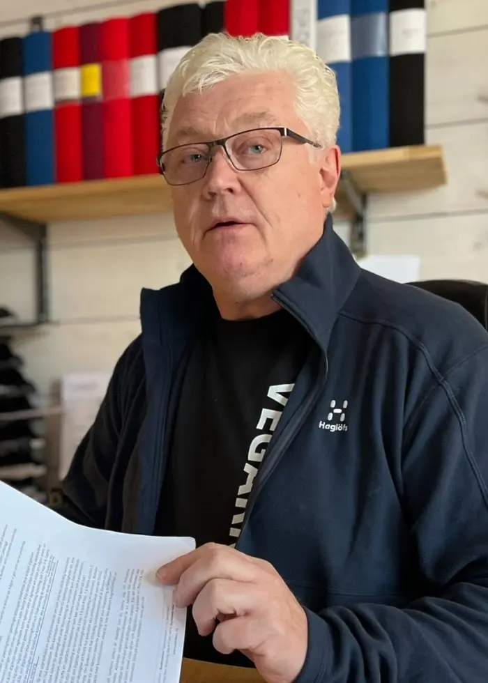 En person sitter vid ett skrivbord med två datorskärmar och håller i papper. Bakom personen finns en hylla med färgkoordinerade pärmar. Arbetsmiljön ser organiserad ut.