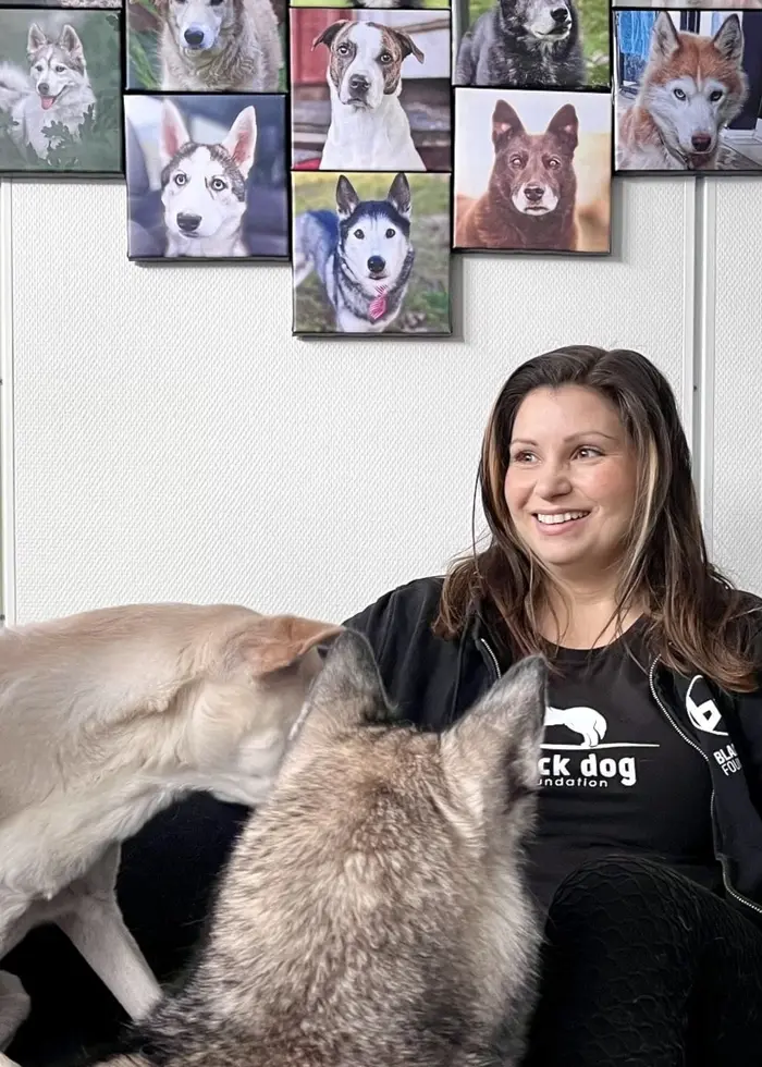 Hundpsykolog Sandra Tigerlind sitter i en soffa omringad av hundar. Bakom henne hänger det massa porträtt av hundar.