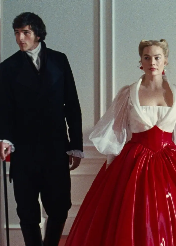 En scen ur filmen ”Wuthering Heights” där två personer står i ett elegant rum. Mannen är klädd i en mörk kostym och håller en käpp, medan kvinnan bär en röd, voluminös klänning med vita ärmar.