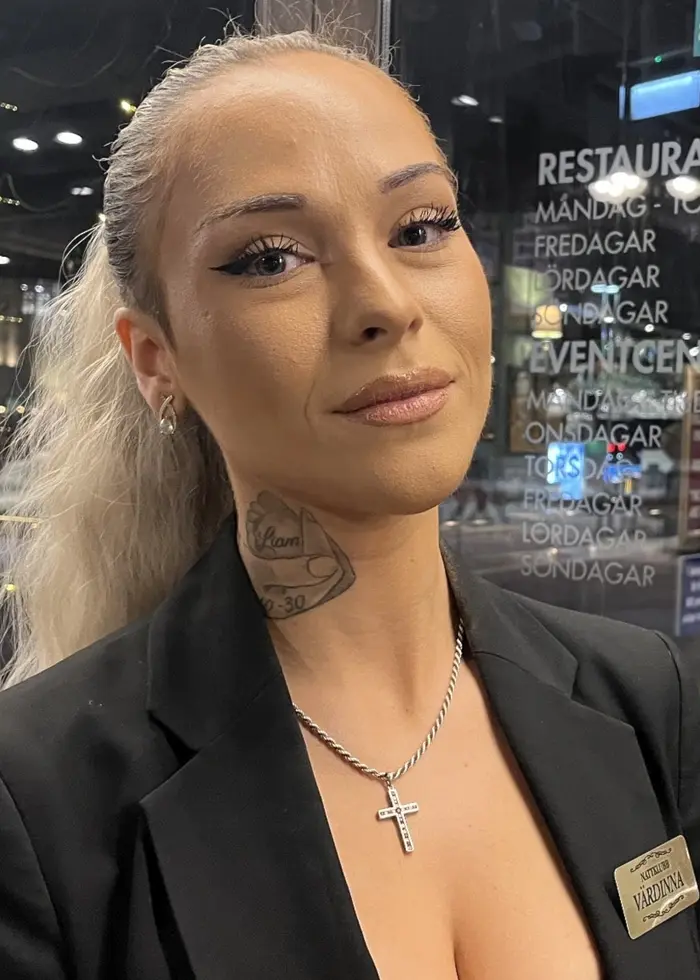 En kvinna med blont hår och en synlig tatuering på halsen står inomhus, klädd i en svart kavaj med ett namnmärke. Bakgrunden visar en restaurangmiljö med ljusslingor och glasfönster.