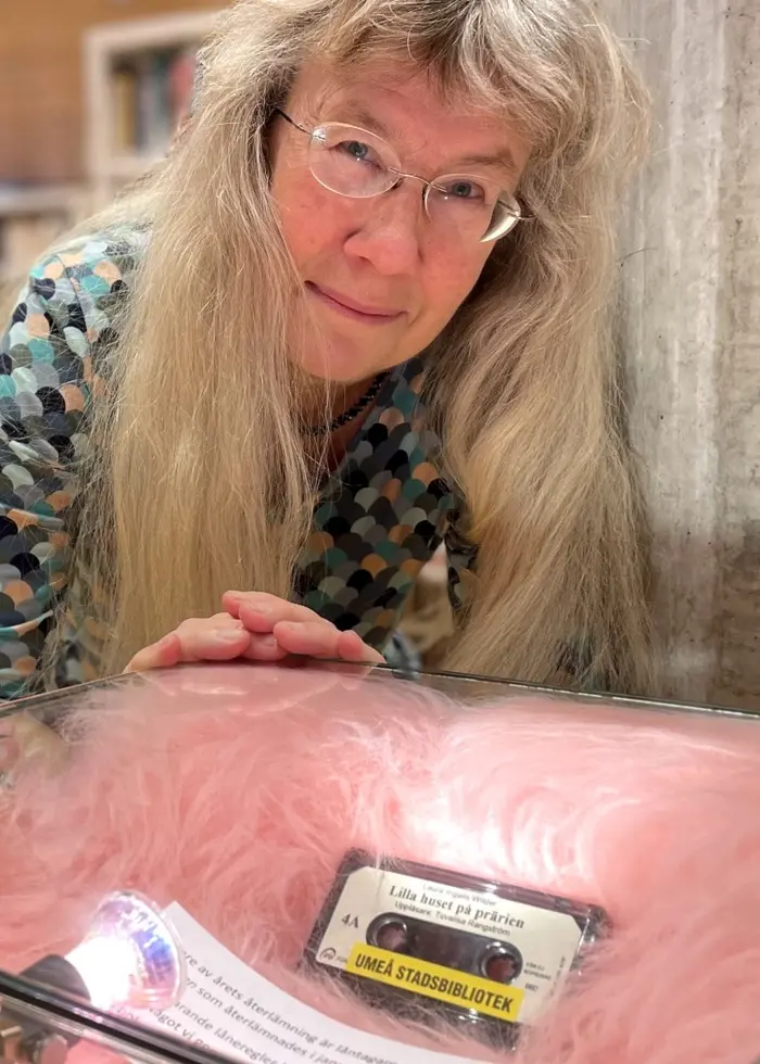 En person med långt blont hår står vid en glasmonter som innehåller ett gult, äldre kassettband och andra föremål. I bakgrunden syns bokhyllor fyllda med böcker i en modern lokal.