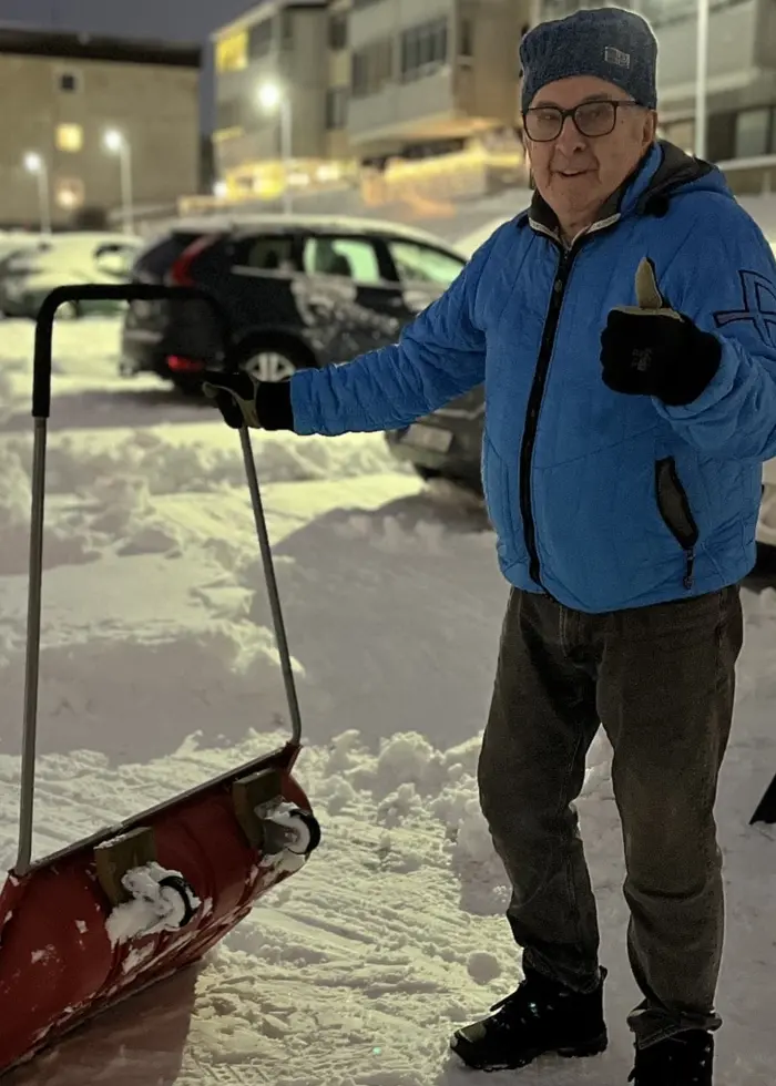 En person klädd i blå jacka och mössa står utomhus på en snötäckt parkeringsplats med en snöskyffel. Bakom personen syns parkerade bilar, bostadshus och en vit skåpbil.