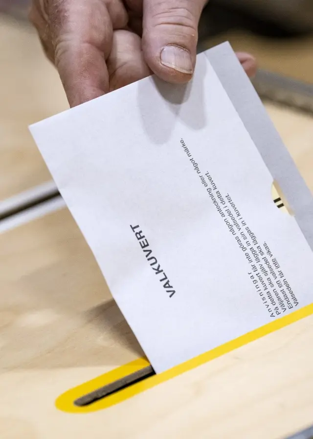 En hand lägger ett valkuvert i en valurna av trä under riksdagsvalet 2022 i Sverige. Bilden visar en guldfärgad öppning på urnan, där kuvertet förs in.