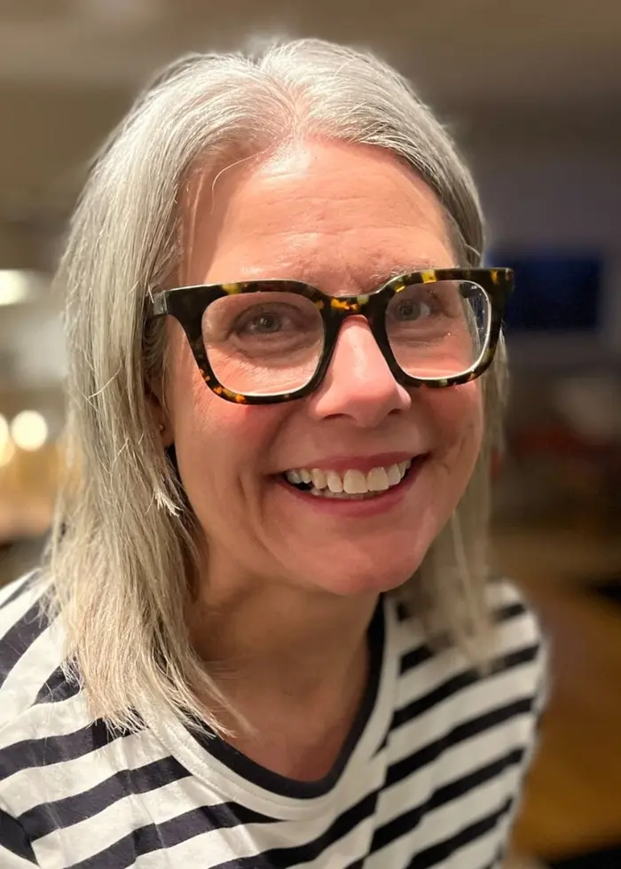 En person med grått hår och randig tröja står i ett hemtrevligt rum med en öppen spis och möbler i bakgrunden. Miljön är mjukt upplyst och ger ett varmt intryck.