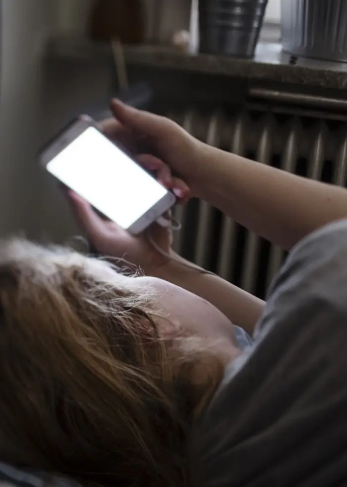 En kvinna ligger i en säng i ett sovrum och håller upp en mobiltelefon. Fönster med gardiner och växter i bakgrunden. Miljön är mörk och avslappnad, med fokus på mobilanvändning.