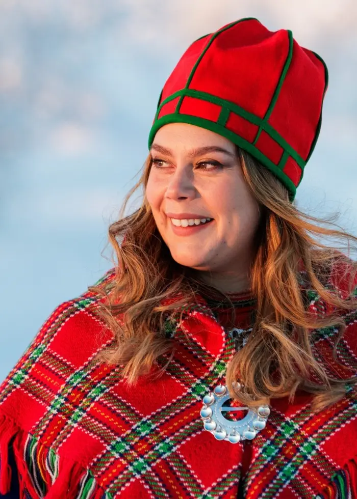 Kvinna i blå sydsamiskkolt, mössa och ullsjal, står i ett vinterlandskap