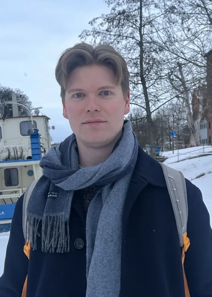 En person står utomhus i ett snöigt landskap i Uppsala, med en båt och träd i bakgrunden. Personen bär en mörk jacka, halsduk och ryggsäck.