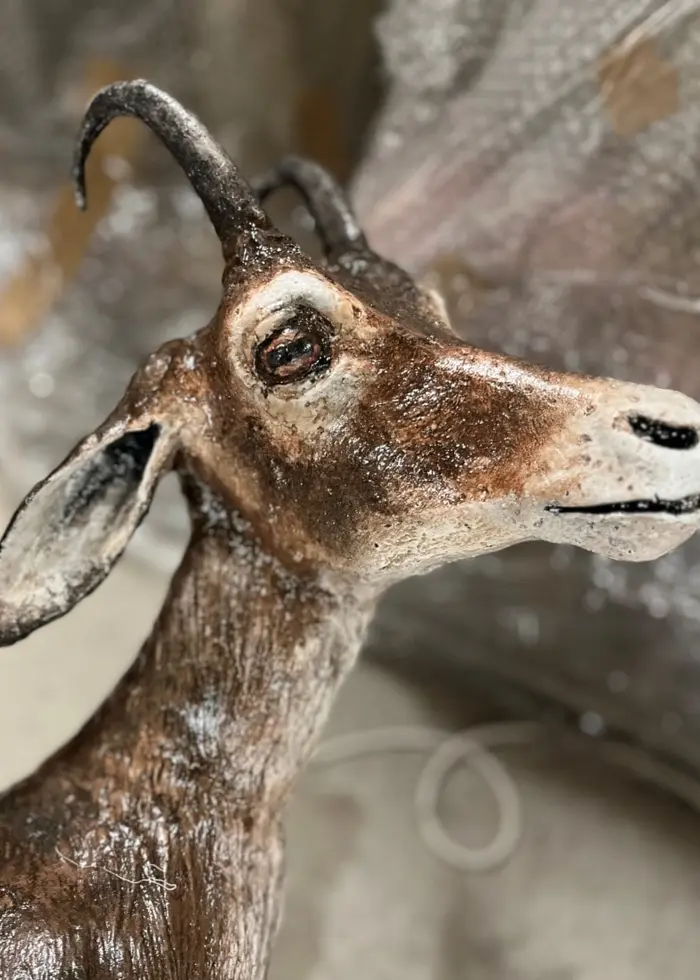 En skäggig man i gul skyddshjälm och reflexjacka på en byggarbetsplats och en huvudet på en bergsget med böjda horn som ingår i ett fasadkonstverk av metall.