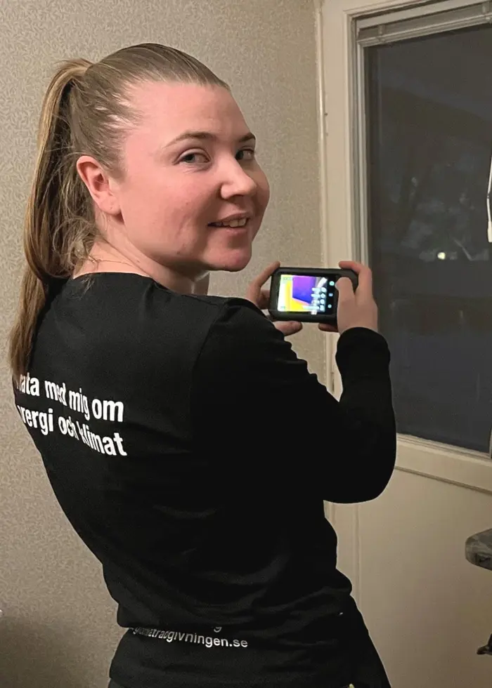 En kvinna i svart tröja står vid ett fönster och håller i en värmekamera. Inomhusmiljön är upplyst och fönstret vetter mot mörker.