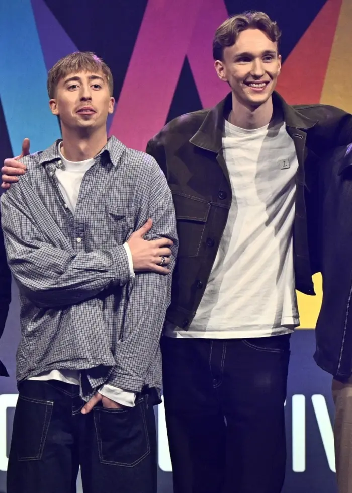 En grupp på fem personer står framför en färgglad Melodifestivalen-logotyp på en scen under en presskonferens. Personerna är klädda i vardagliga kläder och poserar tillsammans för fotografer.