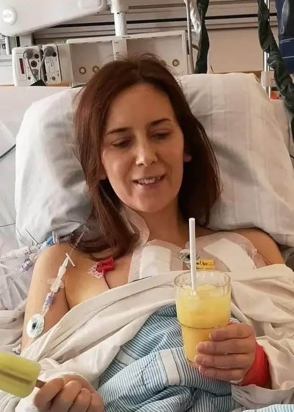 En person ligger i en sjukhussäng omgiven av medicinsk utrustning. Personen är täckt med ett blåvitt täcke och håller i en gul dryck. En monitor syns i bakgrunden.