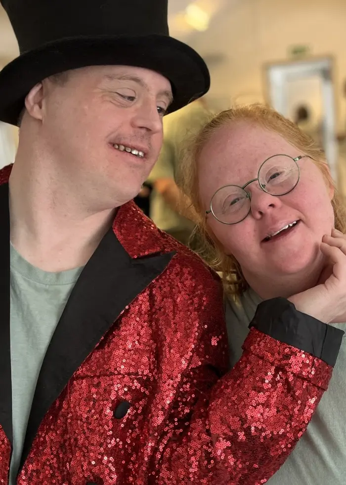 En person i röd paljettkavaj och hög hatt står intill en annan person i ljusgrön tröja. De befinner sig i en ljus inomhusmiljö med flera människor i bakgrunden.