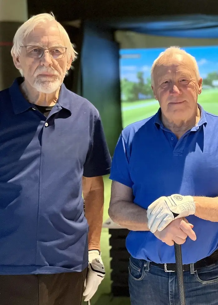 Tre personer står bredvid varandra i en inomhusmiljö med en golfsimulator i bakgrunden. En av dem håller en golfhandske i handen. Alla har mörka kläder, och rummet är upplyst med golftema.