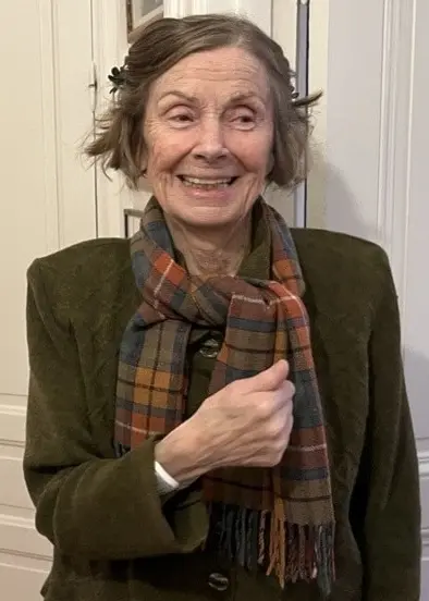 Bilden föreställer en äldre kvinna, 87-åriga Karin, iklädd en mossgrön kavaj med en skotskrutig halsduk som går i mörka färger. Hon har kortklippt hår och står framför två vitmålade garderobsdörrar i en sekelskifteslägenhet. I ena rummet skymtas ett ljust brädgolv och en soffa.