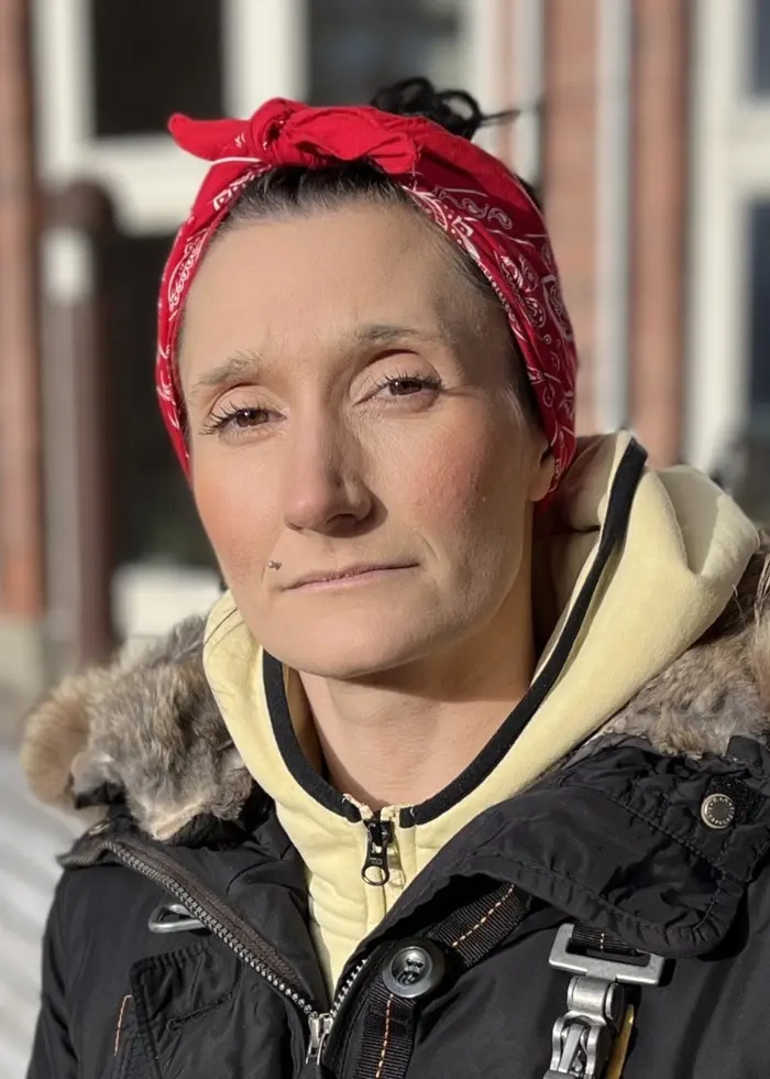 En person med röd bandana och svart jacka med pälsdetaljer står utomhus framför en byggnad med stora fönster. Personen bär en gul huvtröja under jackan.