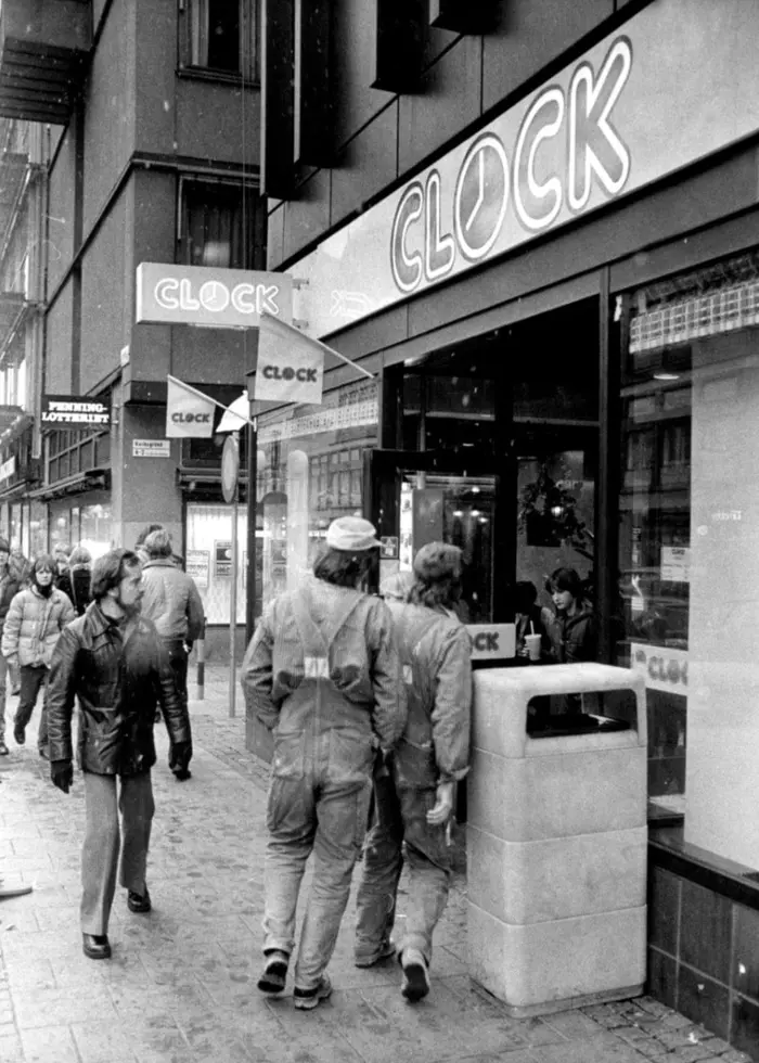 Bilden visar en CLOCK-restaurang på 1970-talet, och människor som går förbi utanför på gatan.