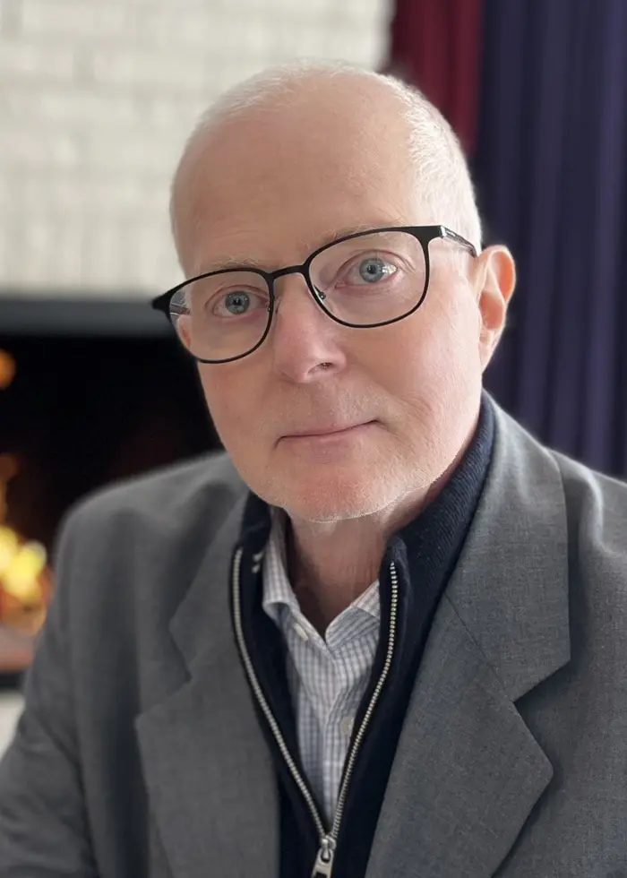 En person i grå kavaj sitter vid ett bord i ett rum med en öppen spis i bakgrunden. Spisen är tänd och en grön växt syns på en hylla till höger.