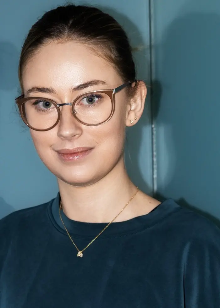 En person står framför en blå vägg, klädd i en mörk tröja och ett halsband. Väggens yta reflekterar personens silhuett tydligt i bakgrunden, vilket skapar en dubbelbildseffekt.