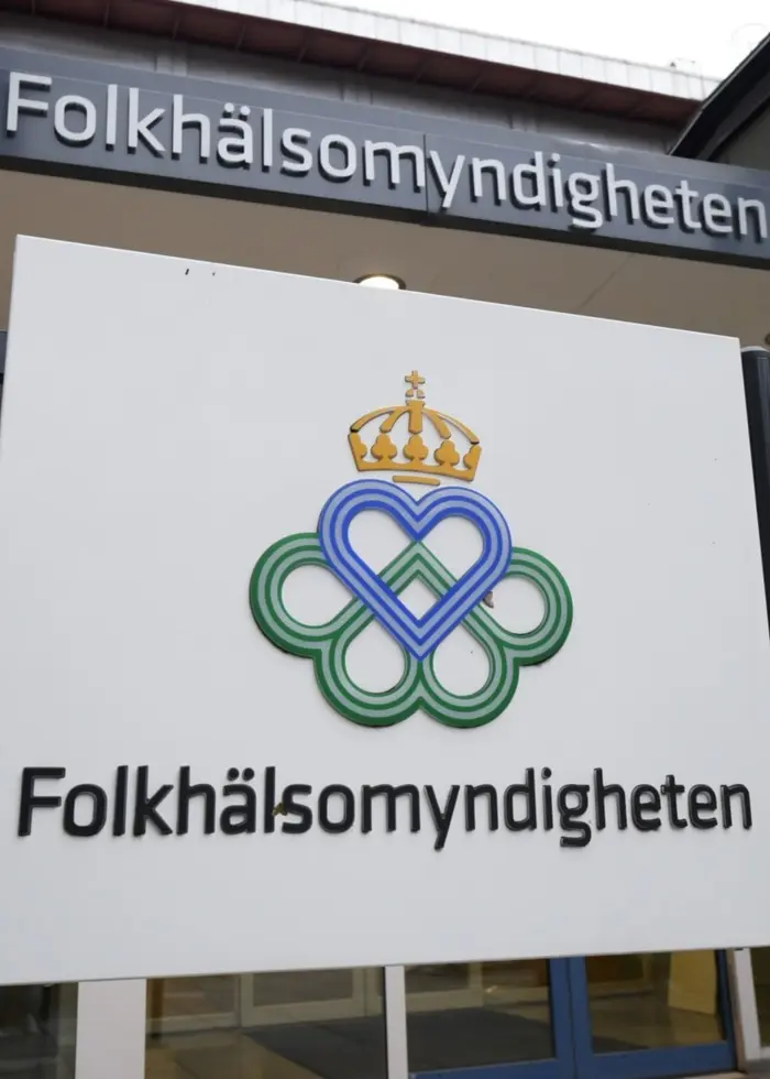 Entrén till Folkhälsomyndighetens byggnad i Solna, Stockholm. En skylt med myndighetens namn och logotyp är synlig framför ingången. Byggnaden har gråa och gröna fasader.