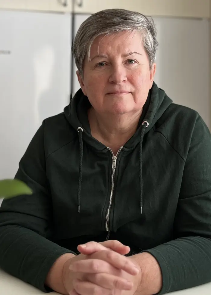 En person sitter vid ett köksbord med händerna knäppta framför sig. Bakgrunden visar ett vitt kylskåp och en vägg med en tavla. Till vänster syns en vas med blommor.