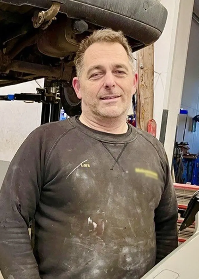 En mekaniker står i en bilverkstad med en bil upplyft bakom honom. Verktyg och reservdelar är spridda på golvet. En annan person arbetar med bilen i bakgrunden.