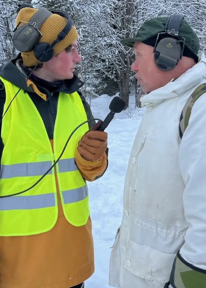 En person i gul reflexväst intervjuar en person i vit vinterjacka och grön mössa på en snötäckt plats med skog i bakgrunden. Fler personer syns längre bort i snön.