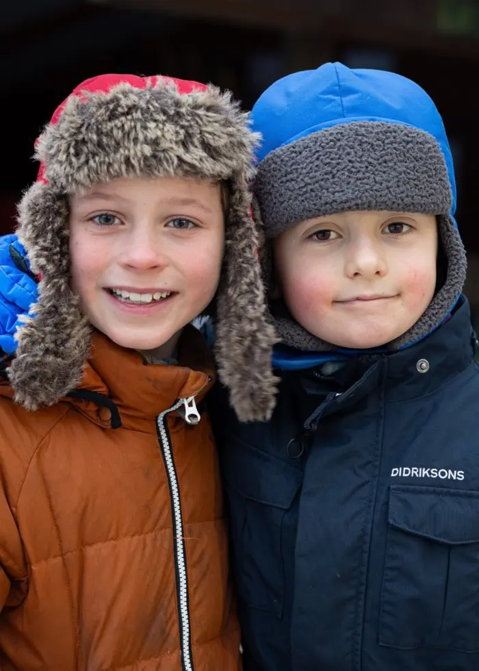 Två barn klädda i varma vinterkläder och pälsmössor står nära varandra utomhus i en snöklädd miljö. En person syns i bakgrunden vid en byggnad.