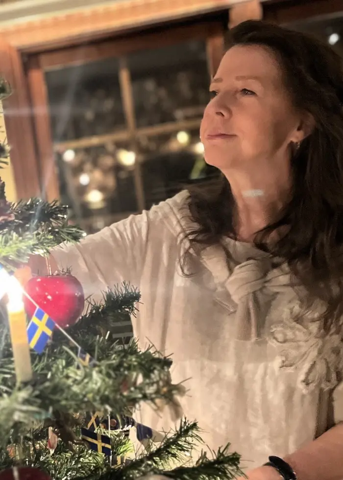 En person klär en julgran med dekorationer, inklusive små svenska flaggor, röda hjärtan och ljus. Granen står i ett rum med fönster och varm belysning.