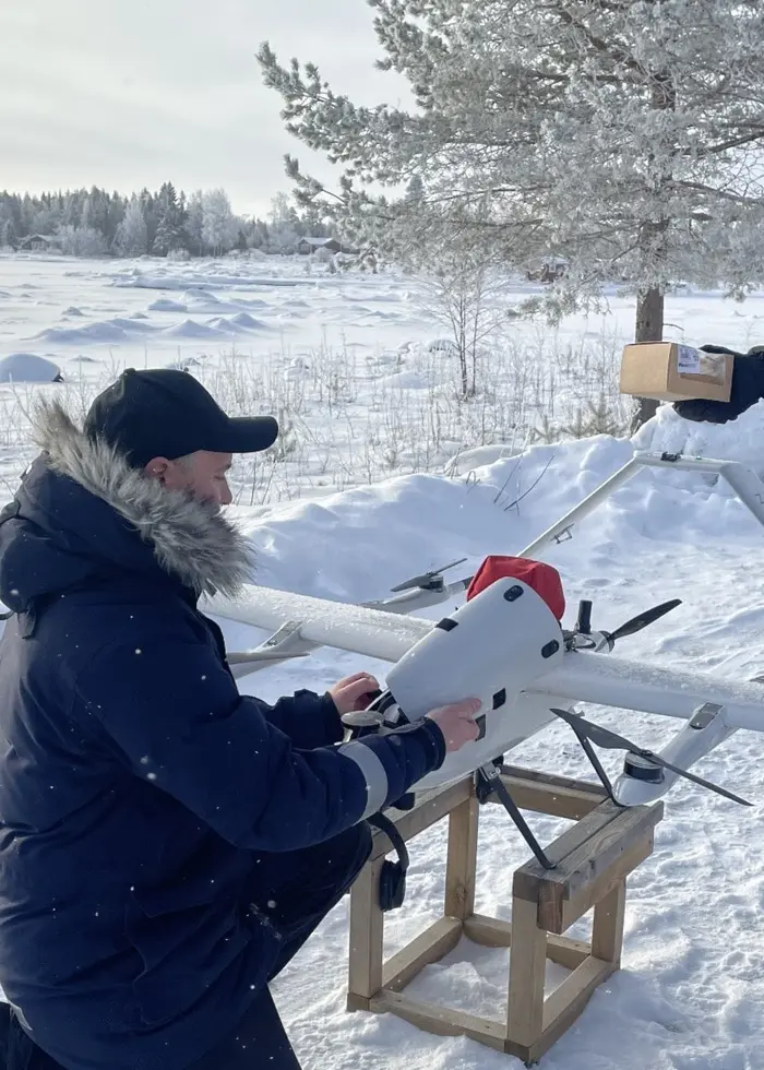 En man i vinterkläder arbetar med en drönare på marken medan en kvinna i lång jacka och mössa står bredvid och håller en låda. De befinner sig i en snöig utomhusmiljö med träd och ett fruset hav i bakgrunden.
