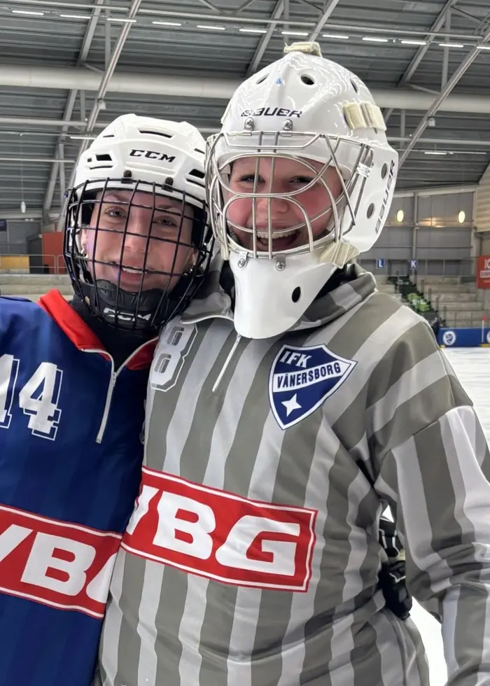 Två bandyspelare står på en isrink inomhus. Den ena bär en blå tröja med röda och vita detaljer och den andra en grå- och vitrandig tröja. Bakom dem syns läktare och arenans takstruktur.